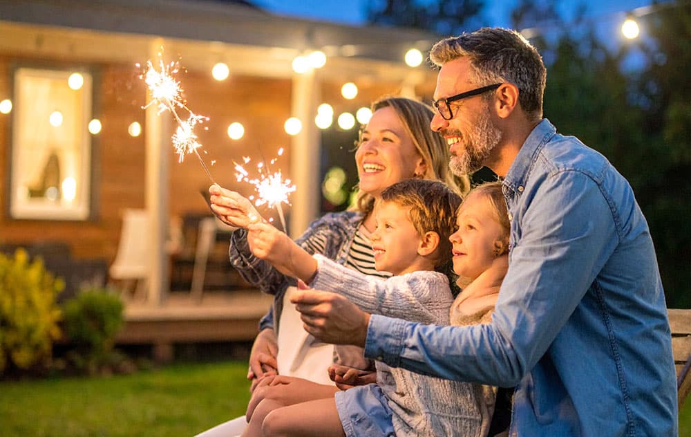 A family watching fireworks from a safe distance in their backyard at night, with one adult wearing protective eyewear while holding a sparkler, promoting fireworks eye safety.