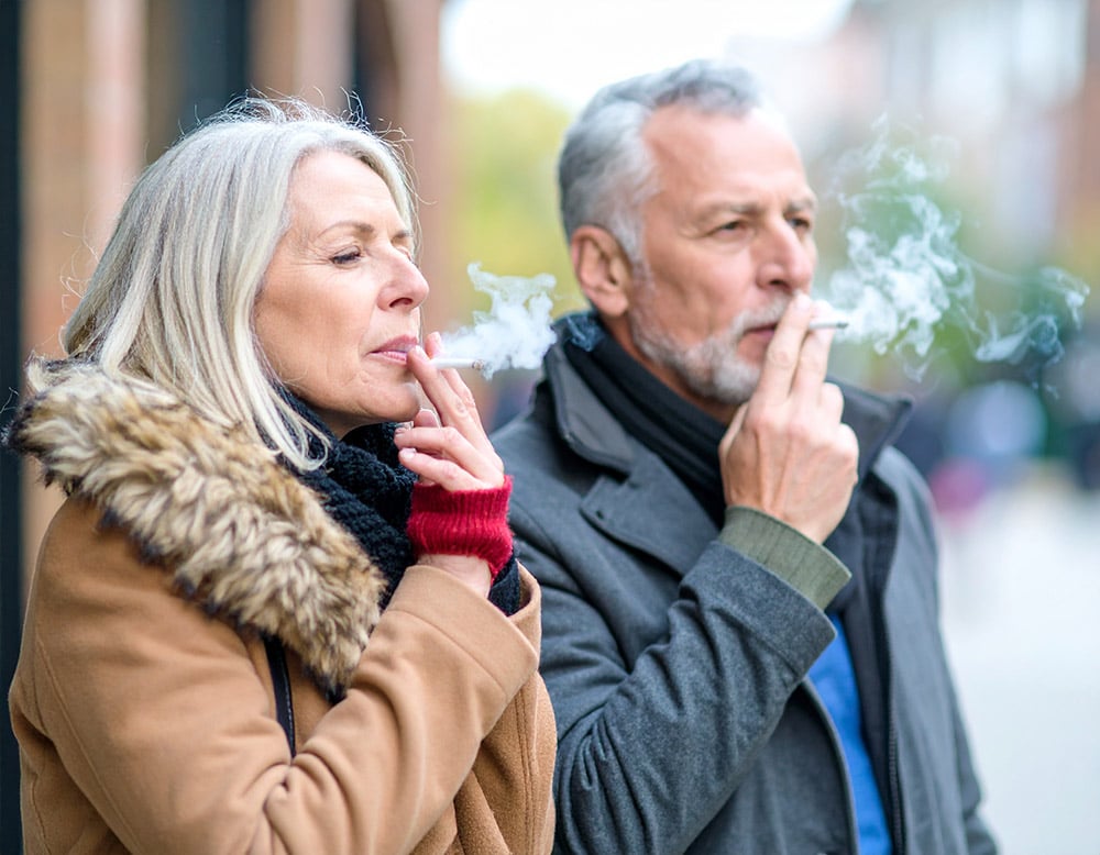 Two adults standing outdoors, casually smoking cigarettes.