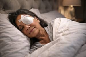 Senior woman sleeping peacefully with an ocular shield during cataract surgery recovery in a low light bedroom setting.