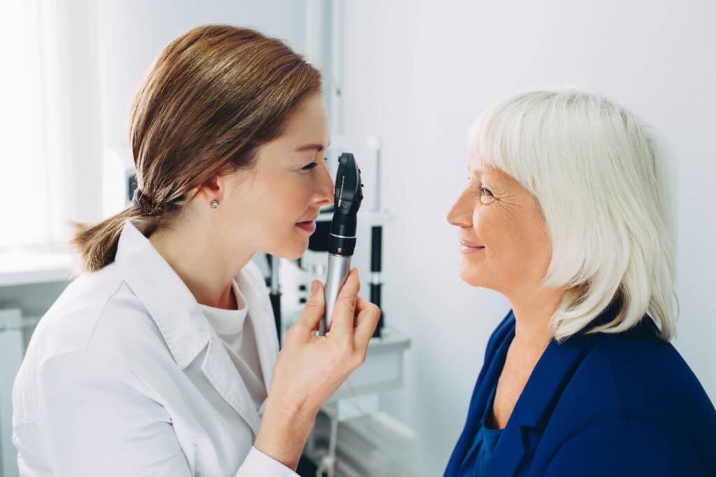 Optometrist holding an ophthalmoscope while examining a smiling patient’s eye.