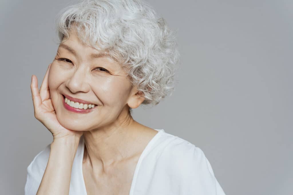 Close-up of a smiling older Asian woman with short gray hair resting her hand on her cheek.