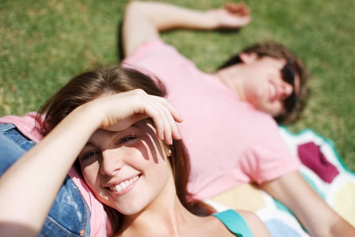 Smiling woman lying on grass and shielding her eyes with her hand, with a man in a pink shirt resting on a blanket in the background.