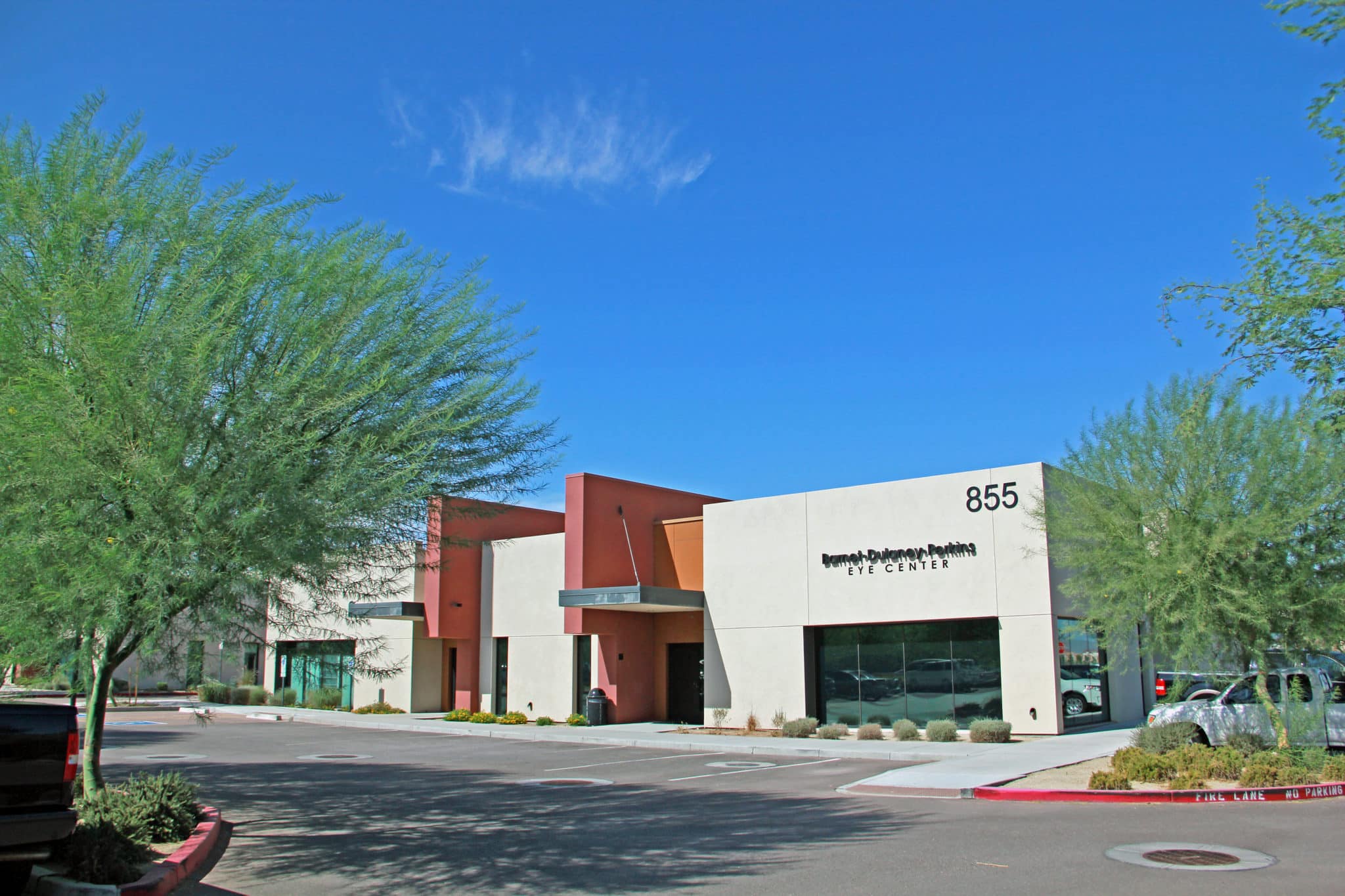Modern, single-story commercial building with a beige and orange facade and the number 855, in a parking lot with trees under a clear blue sky.