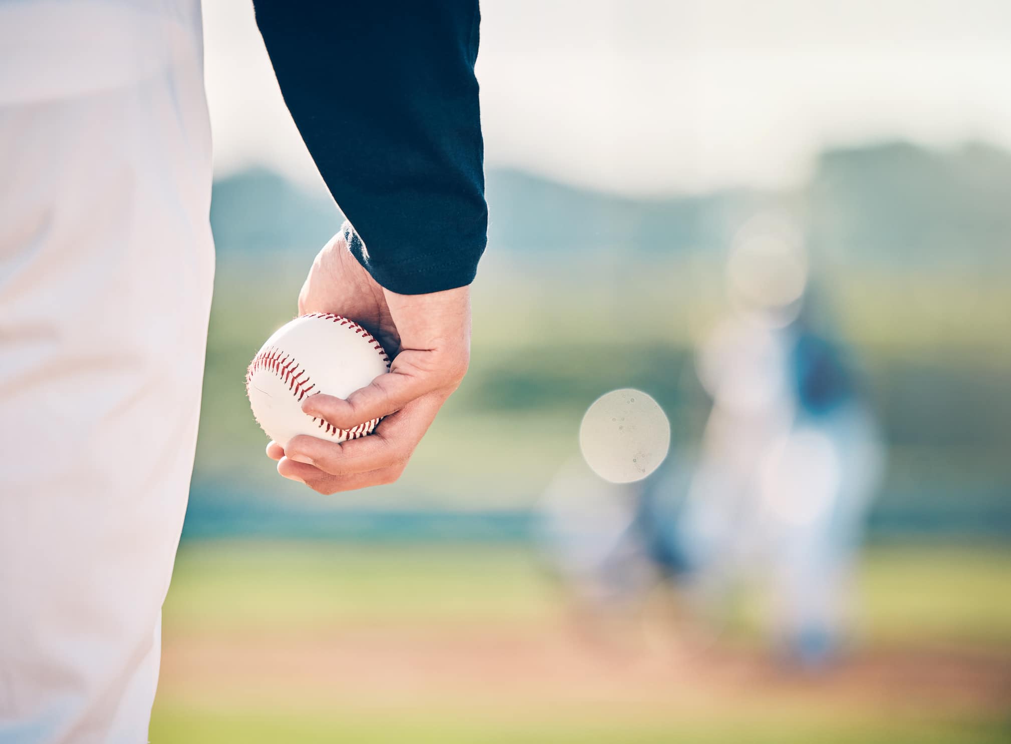 The Science of Seeing and Hitting a Fastball:Close-up of a pitcher’s hand gripping a baseball on the mound, with the batter and catcher blurred in the background.