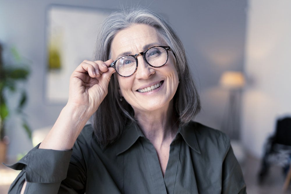 Smiling older woman with gray hair adjusting her eyeglasses, representing improved vision and wondering if she should have cataract surgery.