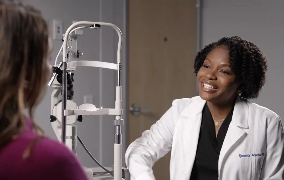 A smiling female doctor in a white coat talks with a patient wearing a purple top in a clinical setting with medical equipment in the background.