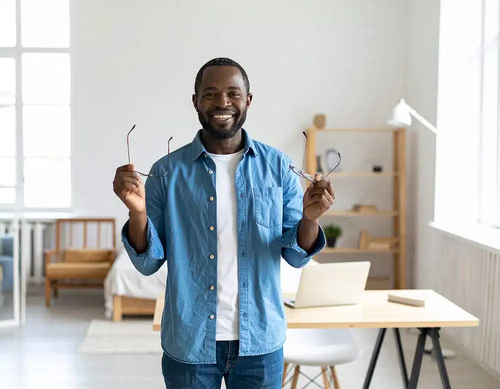Smiling man in a blue shirt stands in a bright, modern office, holding glasses in both hands.