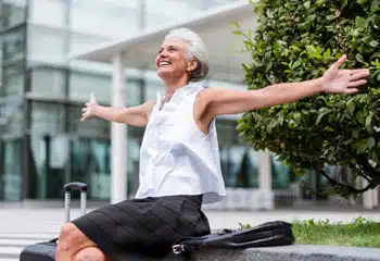 Older woman sitting outdoors with arms open and smiling, representing clearer vision after cataract surgery.