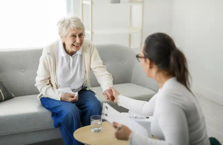 An elderly woman with white hair sits on a sofa, talking to a younger caregiver who is taking notes at a small round table.