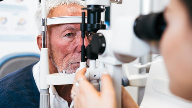 Older male patient receiving an eye exam with a slit-lamp microscope while an eye care professional adjusts the equipment.