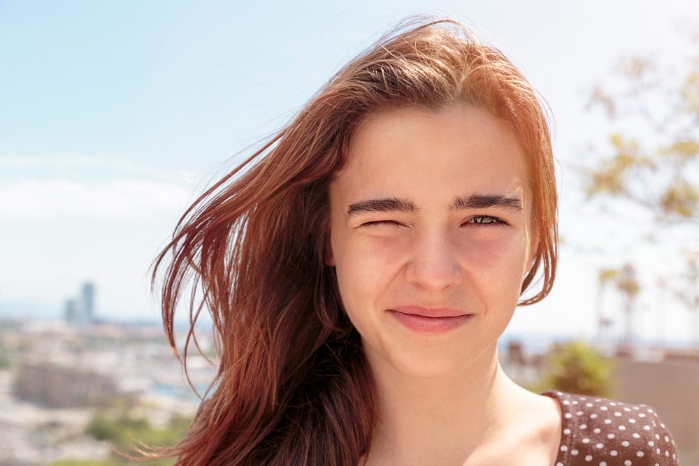 A young woman with wind-blown reddish-brown hair smiles outdoors with a distant blurred cityscape in the background.