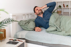 Tired man waking in bed and rubbing one eye, illustrating fatigue as a common trigger for eye twitching.
