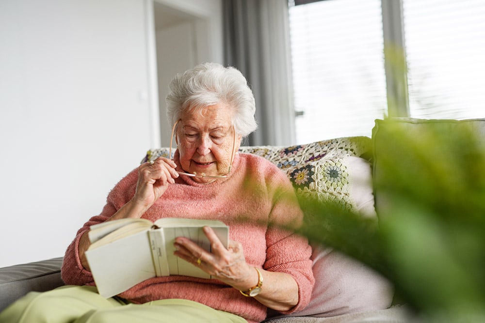 Older woman wearing reading glasses and reading a book at home, illustrating vision changes from a secondary cataract.