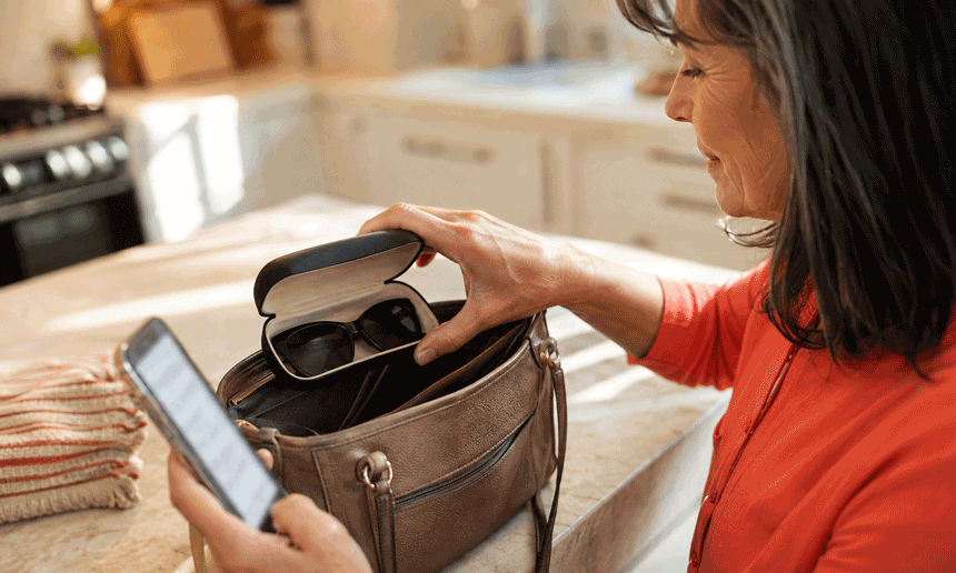 Woman packing sunglasses while checking her phone, illustrating appointment preparation for Barnet Dulaney Perkins Eye Center’s retinal imaging vs dilation guide.